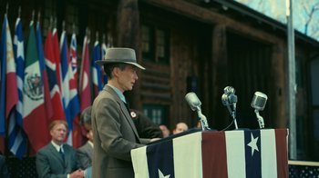 Movie still from “Oppenheimer” (2023), directed by Christopher Nolan – A man in a suit and hat is speaking at a podium; Medium shot, Over the shoulder angle