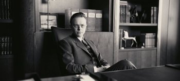 Movie still from “Oppenheimer” (2023), directed by Christopher Nolan – A man in a suit and tie sitting in an office chair; Medium shot, Low angle