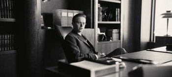 Movie still from “Oppenheimer” (2023), directed by Christopher Nolan – A black and white photo of a man sitting at a desk; Medium shot, Low angle