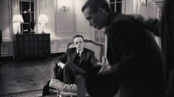 Movie still from “Oppenheimer” (2023), directed by Christopher Nolan – A man in a suit sitting in a chair next to another man in a suit; Medium shot, Low angle