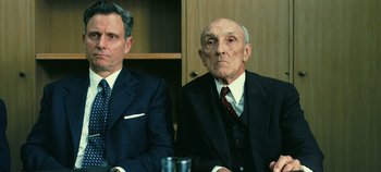 Movie still from “Oppenheimer” (2023), directed by Christopher Nolan – Two men in business suits sitting at a table; Medium shot, Low angle