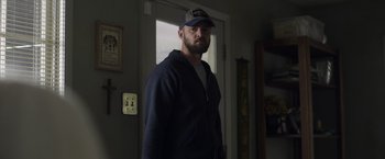 Movie still from “Palmer” (2021), directed by Fisher Stevens – A man wearing a baseball cap standing in front of a door; Medium shot, Low angle