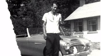 Movie still from “Pamela: A Love Story” (2023), directed by Ryan White – A man standing in front of an old fashioned car; Medium shot, Low angle