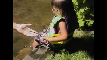 Movie still from “Pamela: A Love Story” (2023), directed by Ryan White – A little girl sitting on a log in the water; Medium shot, Over the shoulder angle
