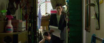 Movie still from “Parasite” (2019), directed by Bong Joon Ho – A man in a suit standing next to a woman in a messy room; Medium shot, Over the shoulder angle
