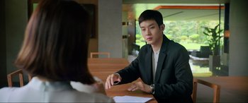 Movie still from “Parasite” (2019), directed by Bong Joon Ho – A man sitting at a table with papers in front of him; Medium shot, Over the shoulder angle