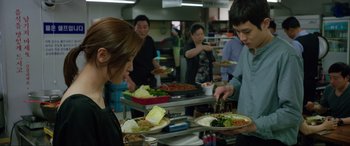Movie still from “Parasite” (2019), directed by Bong Joon Ho – A group of people standing around a table with plates of food on it; Medium shot, Over the shoulder angle