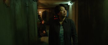 Movie still from “Parasite” (2019), directed by Bong Joon Ho – A woman standing in a room with a man sitting in the background; Medium shot, Over the shoulder angle