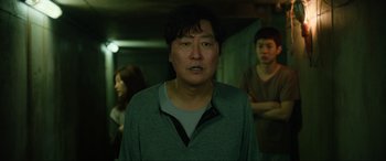 Movie still from “Parasite” (2019), directed by Bong Joon Ho – A man standing in front of two other people; Medium shot, Over the shoulder angle