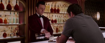 Movie still from “Passengers” (2016), directed by Morten Tyldum – A man in a red jacket pouring a drink into a glass; Medium shot, Over the shoulder angle