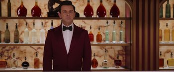 Movie still from “Passengers” (2016), directed by Morten Tyldum – A man in a red jacket and a bow tie in front of bottles; Medium shot, Over the shoulder angle