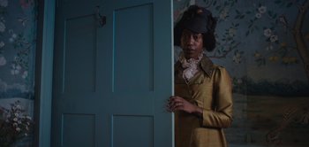 Movie still from “Persuasion” (2022), directed by Carrie Cracknell – A woman standing in front of an open door in a room; Medium shot, Over the shoulder angle