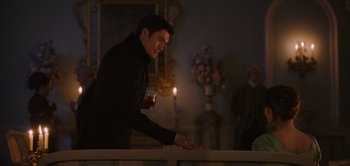 Movie still from “Persuasion” (2022), directed by Carrie Cracknell – A man standing in front of a table holding a candle; Medium shot, Over the shoulder angle
