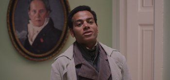 Movie still from “Persuasion” (2022), directed by Carrie Cracknell – A man wearing a suit and tie in front of a painting; Medium shot, Over the shoulder angle
