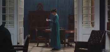 Movie still from “Persuasion” (2022), directed by Carrie Cracknell – A woman in a long dress standing in front of a table; Wide shot, High angle