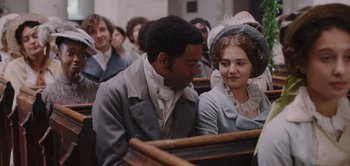 Movie still from “Persuasion” (2022), directed by Carrie Cracknell – A man and a woman sitting in front of each other in front of a crowd; Medium shot, Over the shoulder angle