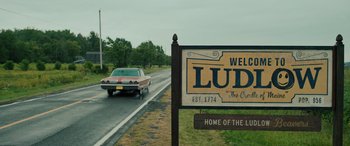 Movie still from “Pet Sematary: Bloodlines” (2023), directed by Lindsey Anderson Beer – A car driving down a road next to a welcome sign; Extreme Wide shot, Low angle
