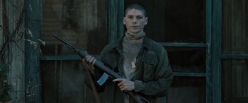Movie still from “Pet Sematary: Bloodlines” (2023), directed by Lindsey Anderson Beer – A young man holding a rifle in front of a window; Medium shot, Low angle
