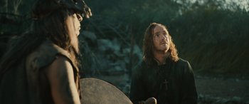 Movie still from “Pet Sematary: Bloodlines” (2023), directed by Lindsey Anderson Beer – A man with long hair and a cross on his chest is standing next to another man with long hair and a cross on his chest; Medium shot, Over the shoulder angle