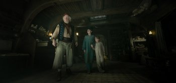 Movie still from “Peter Pan & Wendy” (2023), directed by David Lowery – Two men and a boy are standing in a dark room; Wide shot, Low angle