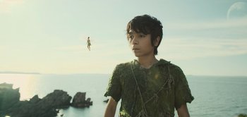 Movie still from “Peter Pan & Wendy” (2023), directed by David Lowery – A young boy wearing a green shirt standing in front of a body of water; Medium shot, Low angle