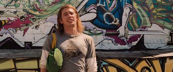 Movie still from “Pineapple Express” (2008), directed by David Gordon Green – A man with long blonde hair standing in front of a mural; Medium shot, Low angle
