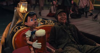 Movie still from “Pinocchio” (2022), directed by Robert Zemeckis – A man sitting next to a statue of a boy holding a glass of beer; Medium shot, Over the shoulder angle