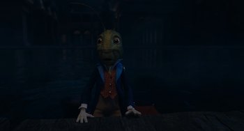 Movie still from “Pinocchio” (2022), directed by Robert Zemeckis – A creepy looking creature sitting on top of a wooden table; Medium shot, High angle