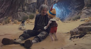 Movie still from “Pinocchio” (2022), directed by Robert Zemeckis – A man sitting on the ground next to a puppet; Wide shot, High angle
