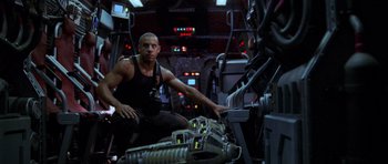 Movie still from “Pitch Black” (2000), directed by David Twohy – A man sitting on a chair next to another man in a room; Medium shot, Low angle