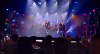 Movie still from “Pitch Perfect” (2012), directed by Jason Moore – A group of people standing on a stage in front of lights; Extreme Wide shot, Over the shoulder angle