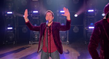 Movie still from “Pitch Perfect” (2012), directed by Jason Moore – A man standing on a stage holding a microphone; Medium shot, Low angle