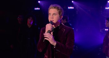 Movie still from “Pitch Perfect” (2012), directed by Jason Moore – A young man holding a microphone while standing in front of a group of people; Medium shot, Over the shoulder angle