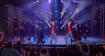 Movie still from “Pitch Perfect” (2012), directed by Jason Moore – A group of people on a stage with lights in the background; Extreme Wide shot, Overhead angle