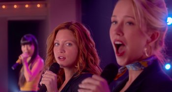 Movie still from “Pitch Perfect” (2012), directed by Jason Moore – Two women sing into microphones in front of purple lights; Close Up shot, Over the shoulder angle