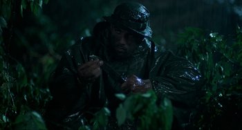 Movie still from “Platoon” (1986), directed by Oliver Stone – A man in a rain jacket and a rain hat is in the woods; Medium shot, Low angle