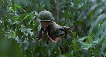 Movie still from “Platoon” (1986), directed by Oliver Stone – A man in a helmet is holding a rifle in the woods; Medium shot, Low angle