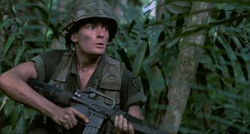 Movie still from “Platoon” (1986), directed by Oliver Stone – A soldier holding an m - 1 6 rifle while standing next to a tree; Close Up shot, Low angle