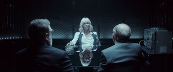 Movie still from “Atomic Blonde” (2017), directed by David Leitch – A woman sitting in front of two men in front of a mirror; Medium shot, Over the shoulder angle