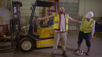 Movie still from “Popovich: Road to Hollywood” (2021), directed by Gregory Popovich – A man with a beard and a yellow vest standing in front of a forklift; Wide shot, Low angle