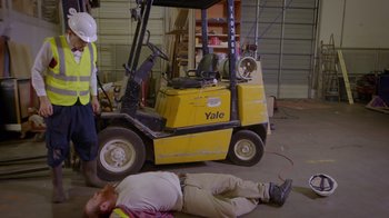 Movie still from “Popovich: Road to Hollywood” (2021), directed by Gregory Popovich – A man laying on the ground in front of a forklift; Wide shot, High angle