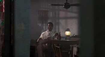 Movie still from “Predator” (1987), directed by John McTiernan – A man sitting at a table in a room with a lamp; Wide shot, Low angle