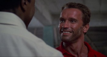 Movie still from “Predator” (1987), directed by John McTiernan – A person smiling at a person; Close Up shot, Over the shoulder angle