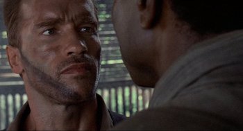 Movie still from “Predator” (1987), directed by John McTiernan – Two men are facing each other in front of a window; Close Up shot, Over the shoulder angle
