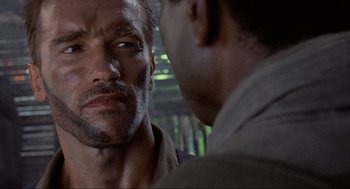 Movie still from “Predator” (1987), directed by John McTiernan – A man looking at another man in a dark room; Close Up shot, Over the shoulder angle