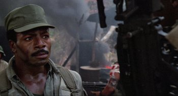 Movie still from “Predator” (1987), directed by John McTiernan – A man in a green hat is standing in front of a fire; Close Up shot, Over the shoulder angle