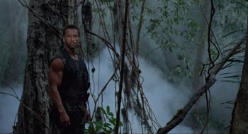 Movie still from “Predator” (1987), directed by John McTiernan – A man standing next to a tree in the woods; Medium shot, Low angle