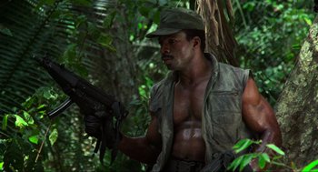 Movie still from “Predator” (1987), directed by John McTiernan – A man holding a gun while standing next to a tree; Medium shot, Low angle