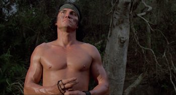 Movie still from “Predator” (1987), directed by John McTiernan – A shirtless man standing in front of a tree; Medium shot, Low angle