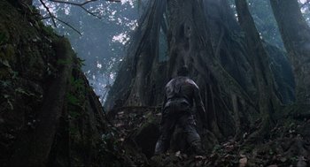 Movie still from “Predator” (1987), directed by John McTiernan – A man standing next to a tree in the woods; Wide shot, Low angle
